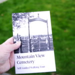 Image Description: A close up of a left hand holding a Mountain View Cemetery Self Guided Walking Tour Booklet. The booklet cover has a title and an image of the old Jewish Cemetery gates. The Cemetery grounds are out of focus in the background of the photo.