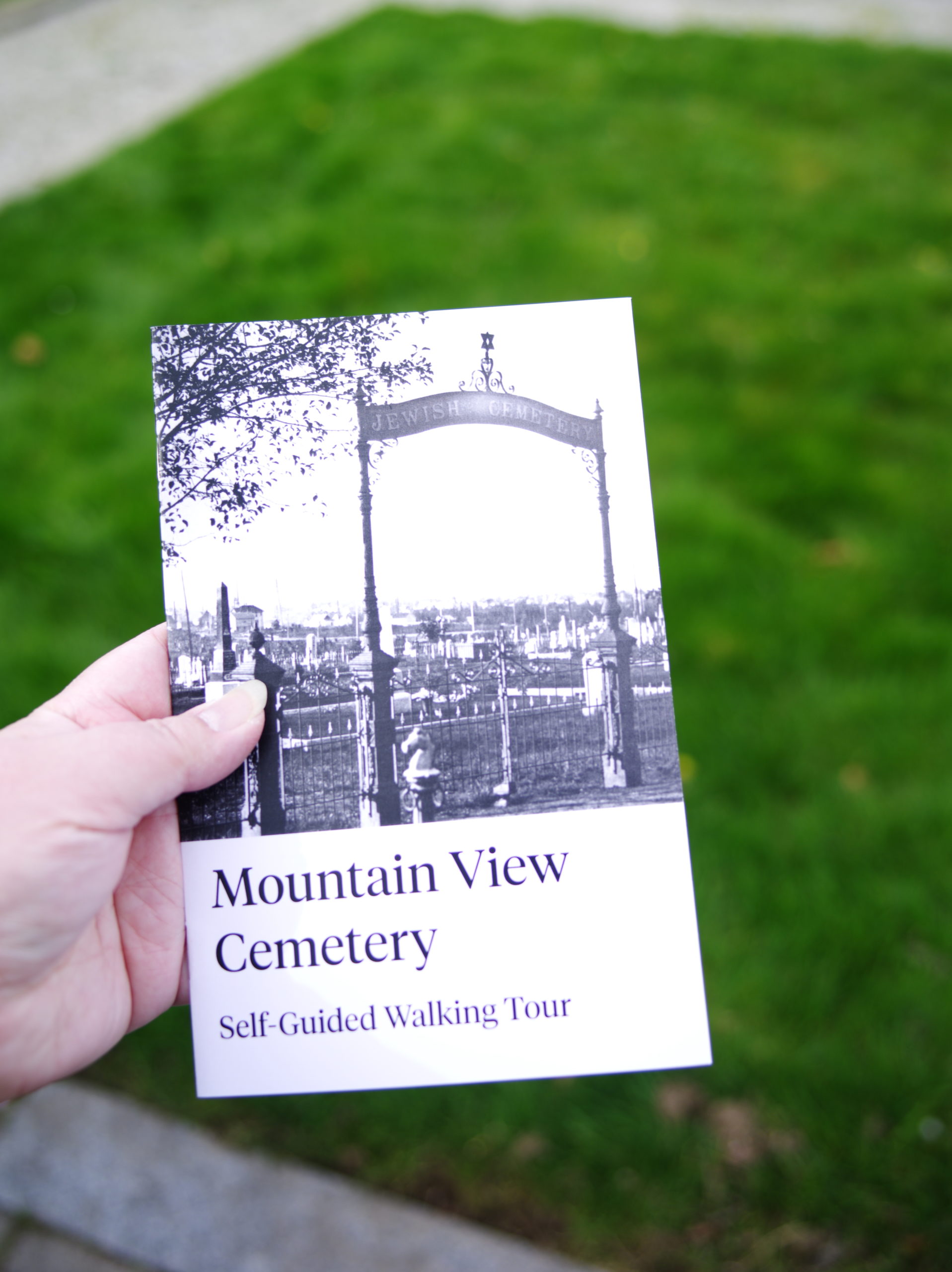 Image Description: A close up of a left hand holding a Mountain View Cemetery Self Guided Walking Tour Booklet. The booklet cover has a title and an image of the old Jewish Cemetery gates. The Cemetery grounds are out of focus in the background of the photo.