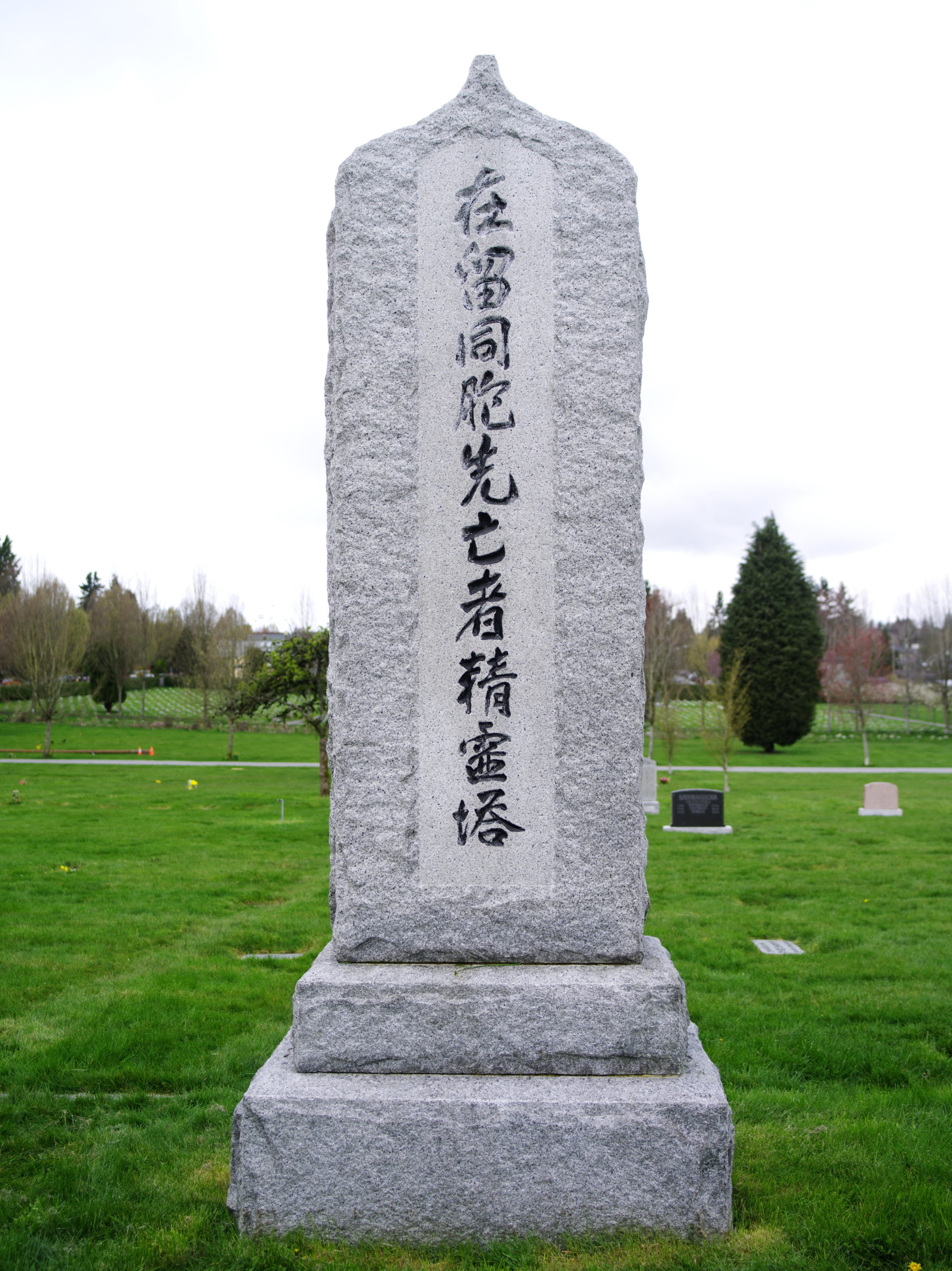 Image Description: A tall, narrow headstone is centered in the photograph. It sits on two square bases. There is an inscription, written vertically, in mixed scripts of Kanji (read nihongo) and Kana (Hiragana, Katakana). The inscription is within a smoothed area on the surface of the headstone. The inscription characters (text) are black.