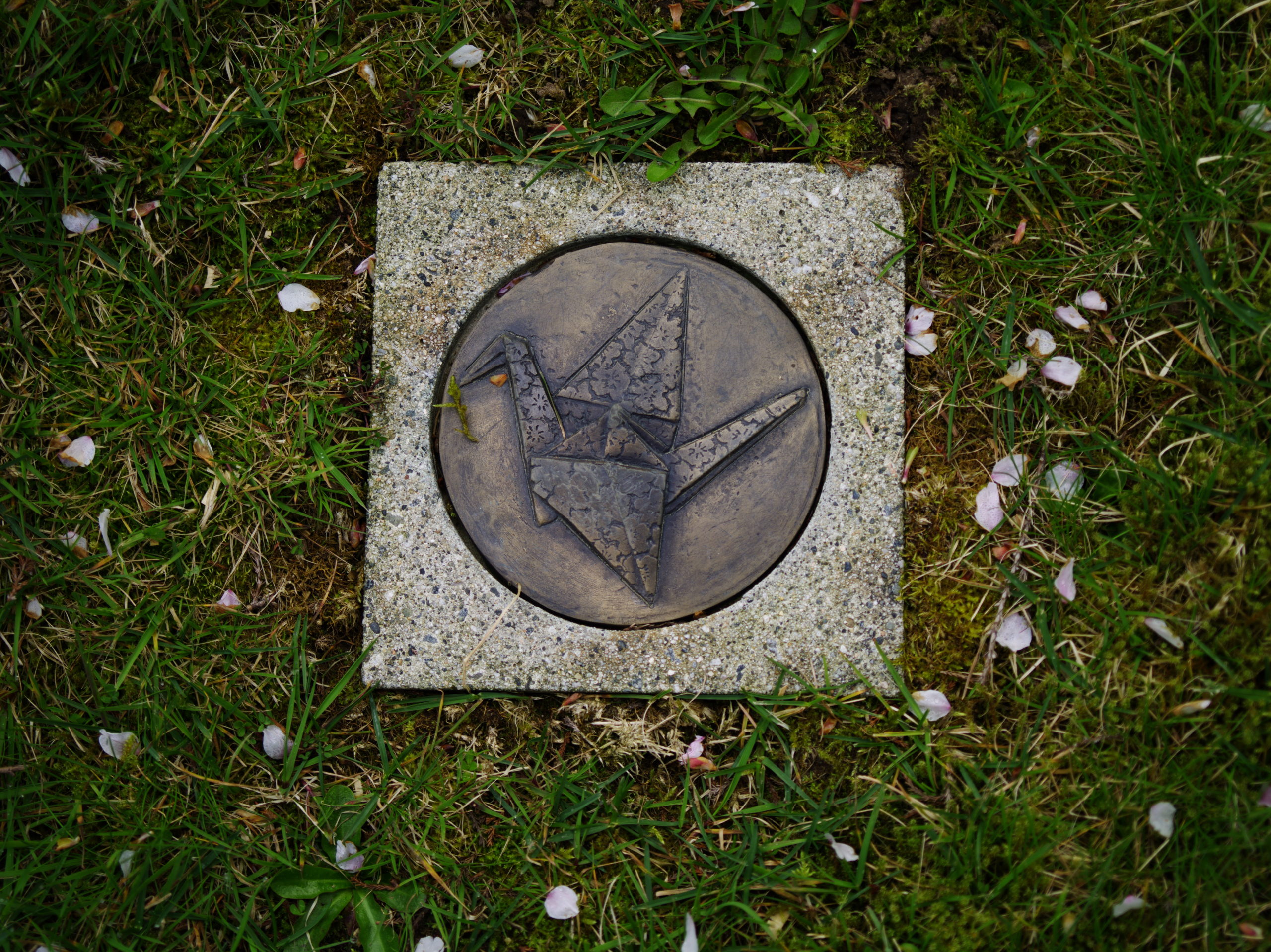 Image Description: A square flat stone frames a round circle featuring an image of an origami crane. There are cherry blossom petals on the grass that surround this small commemorative marker.The crane is designed with many small cherry blossoms patterned over its body.