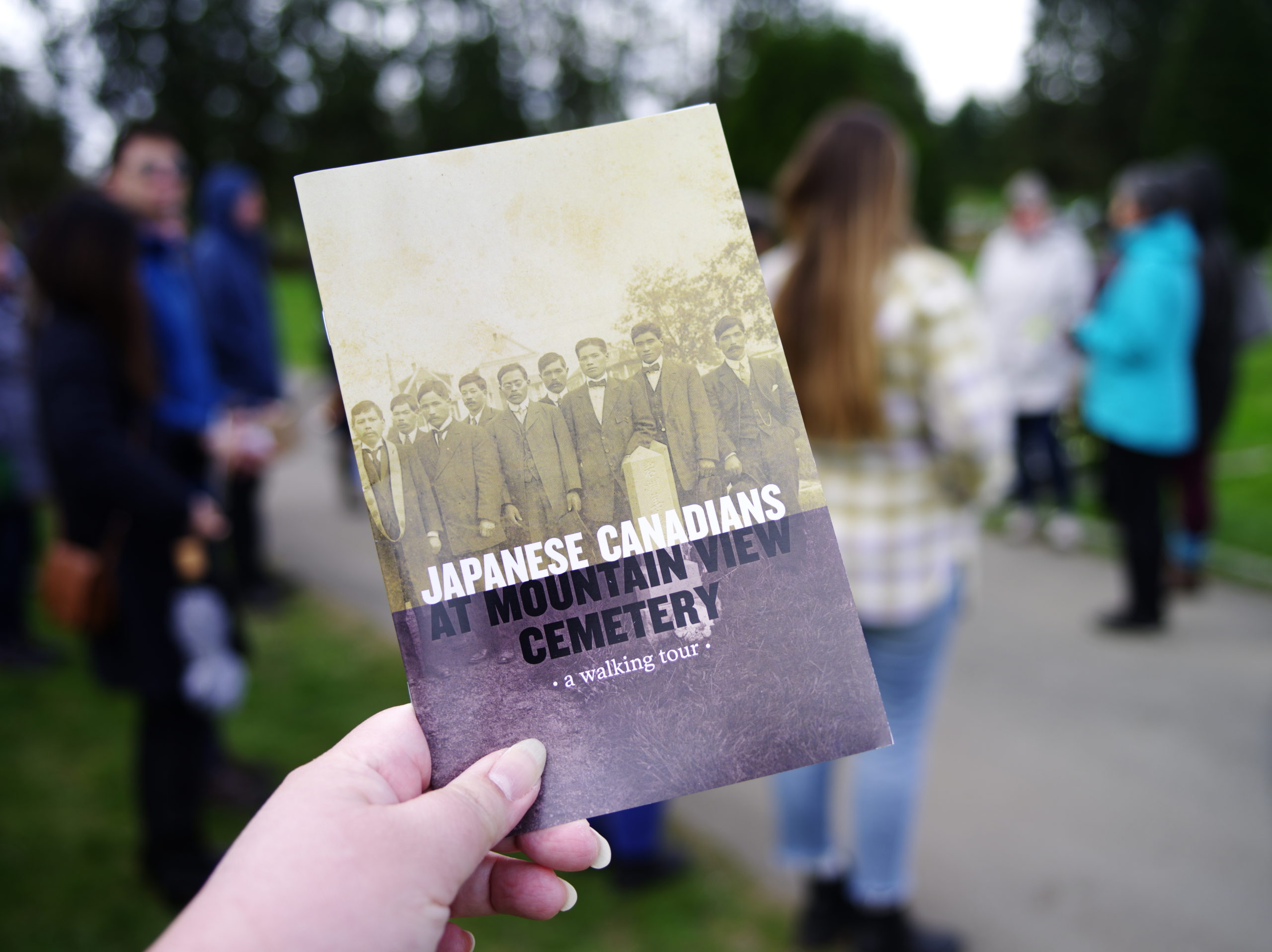 Image Description: A close up photo of a left hand holding a brochure. The sepia toned black and white cover image of the brochure has nine people standing in a straight line. They are wearing western style suit jackets, vests, and ties. The written English text on the cover over the legs of the standing people reads: Japanese Canadiens At Mountain View Cemetery a walking tour