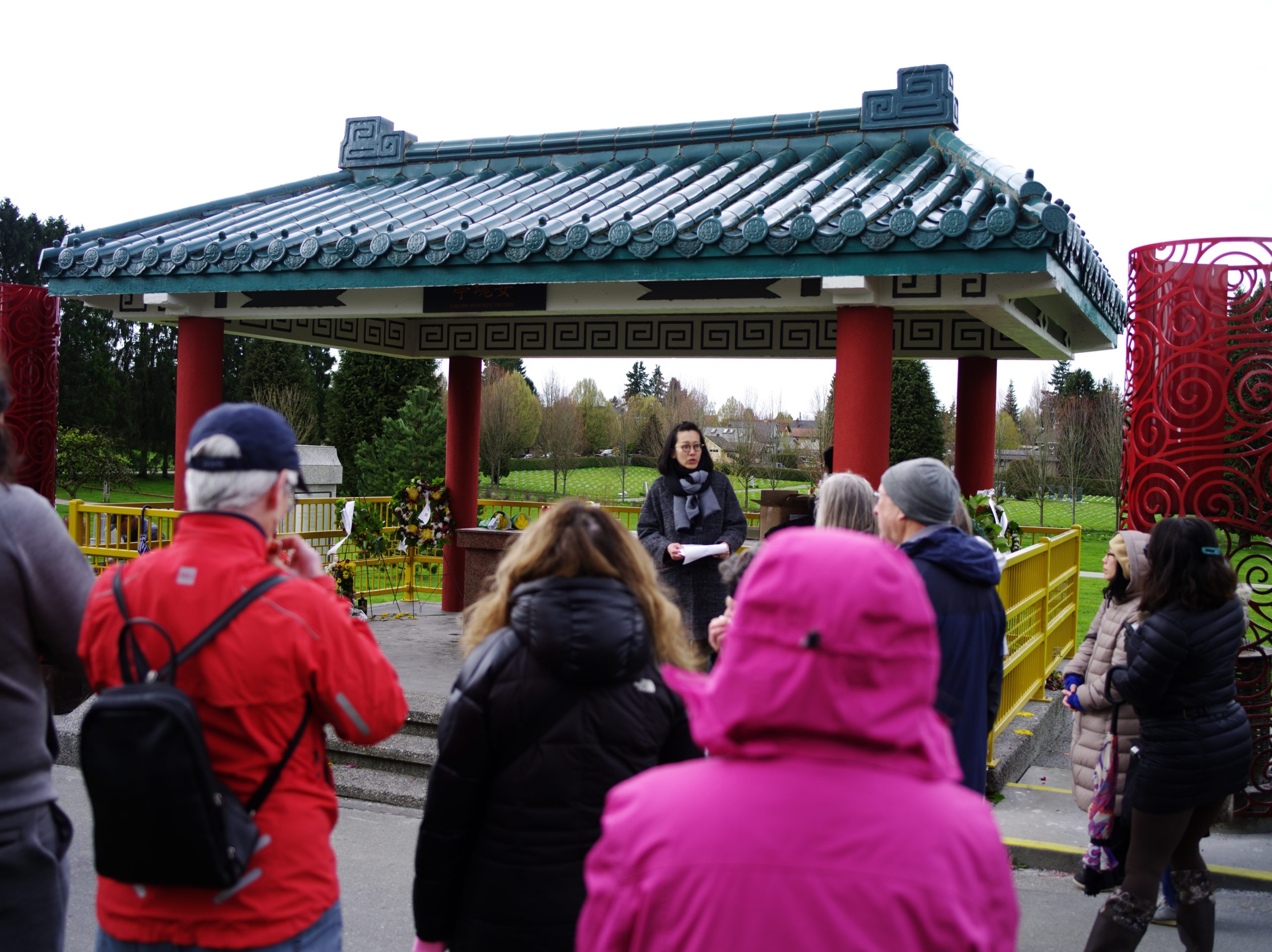 Image Description: A person stands in the north east corner of the Chinese Pavilion at Mountain View Cemetery. They have shoulder length black hair, with their bangs pulled back away from their face. They are reading welcome notes from a notepad they are holding in their hands. The pavilion roof is held up by four round brightly painted pillars. There are black and white designs on the inside of the roof. Shiny, decorative cylindrical burners featuring a round swirly pattern are on either side of the pavilion. People are facing the pavilion, looking at the person holding the notepad.