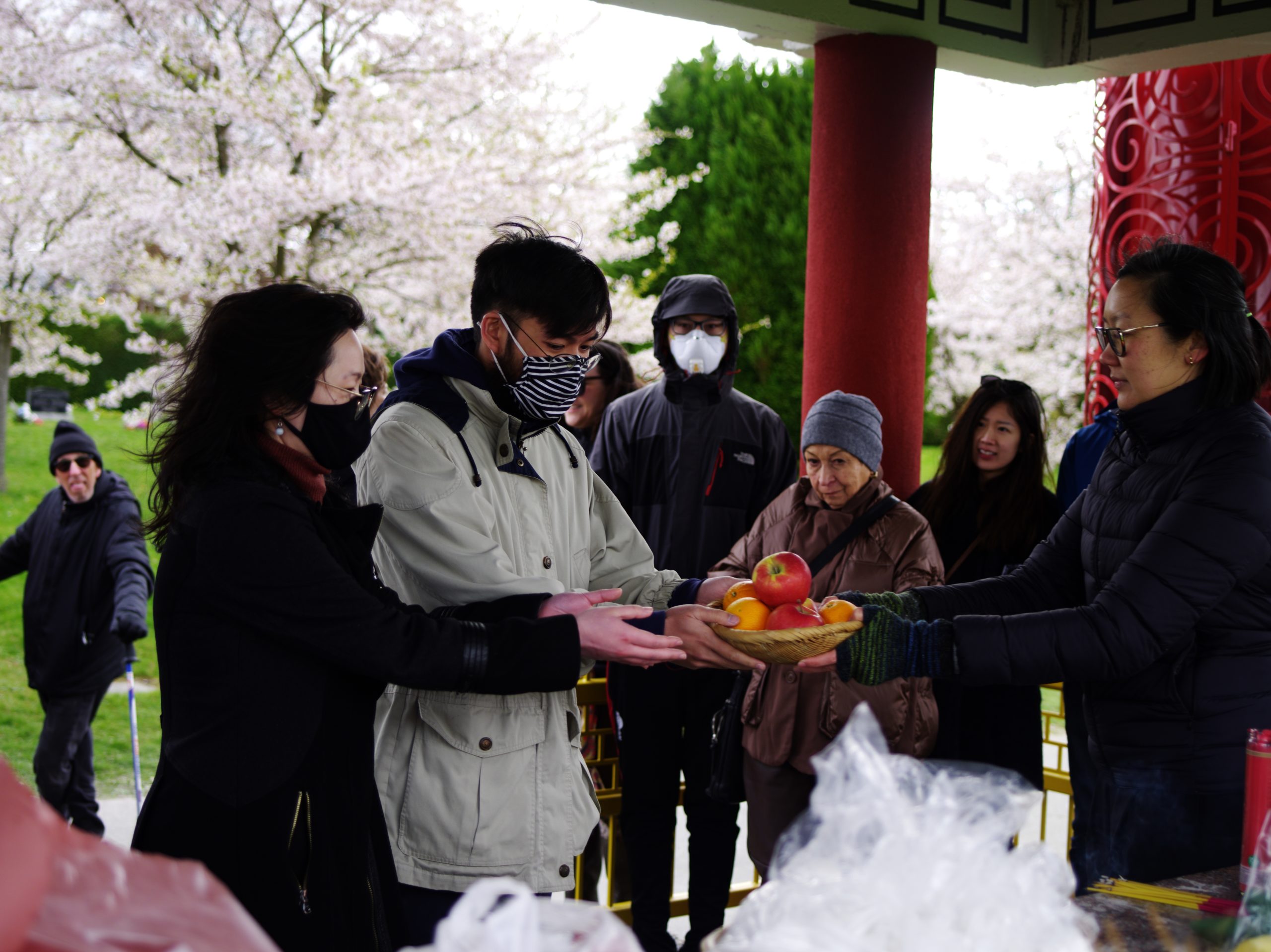 Image Description:Three people stand inside the Chinese Pavilion at Mountain View Cemetery. A person with black shoulder length hair and glasses is wearing a puffy jacket. They offer a woven bowl filled with apples and oranges to two other people. The two people stretch their arms out to receive the bowl with their bare hands. Three other people standing at the edge of the pavilion look on at the exchange of gifts. Other gifts for exchange are on the Altar in the foreground of the photograph.