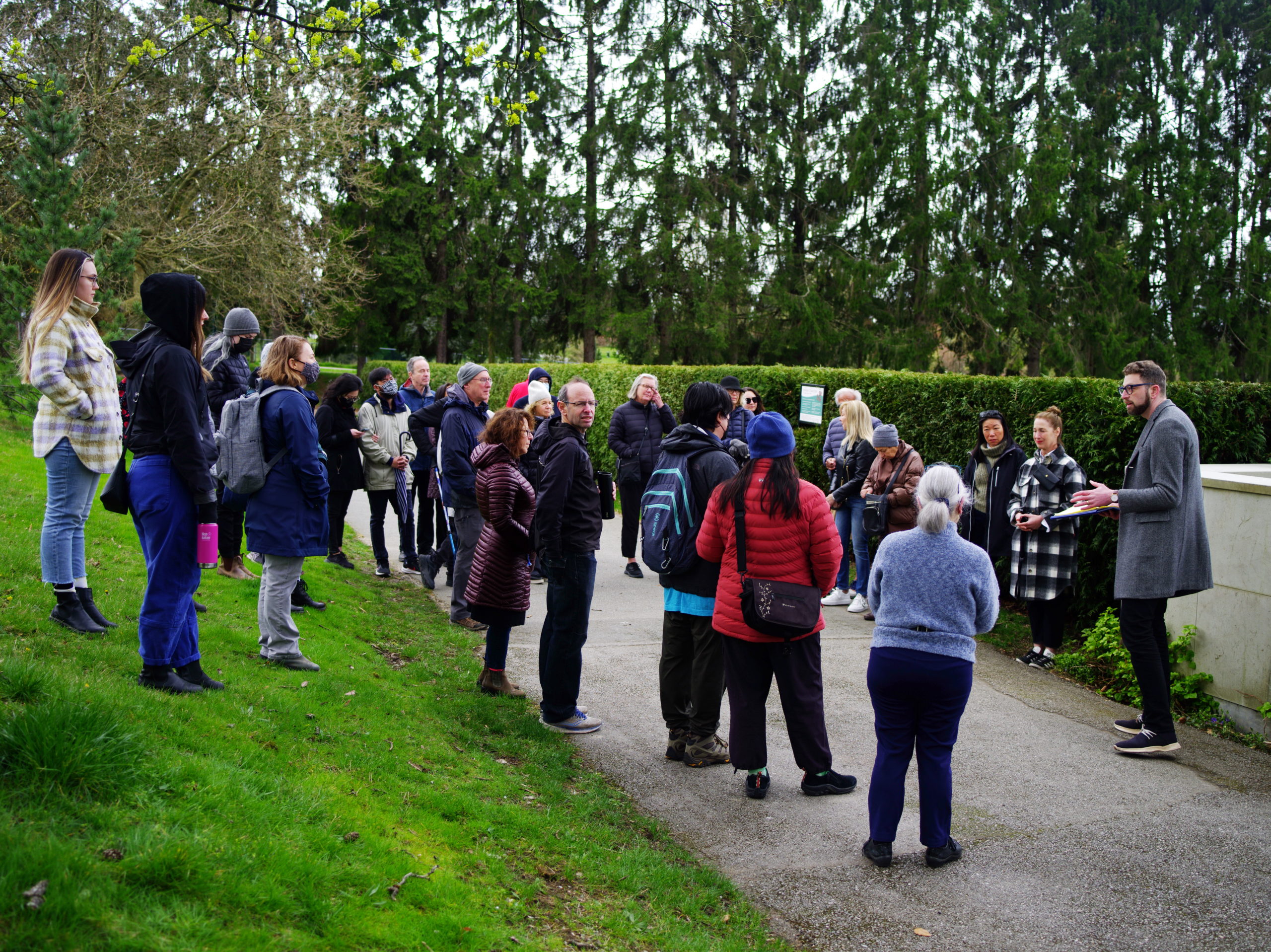 Image Description: Michael Schwartz welcomes a group of people to the Cross Cultural Walking Tours.The people face Michael, standing on a paved path and a grassy hill outside the Jewish Cemetery hedge line.