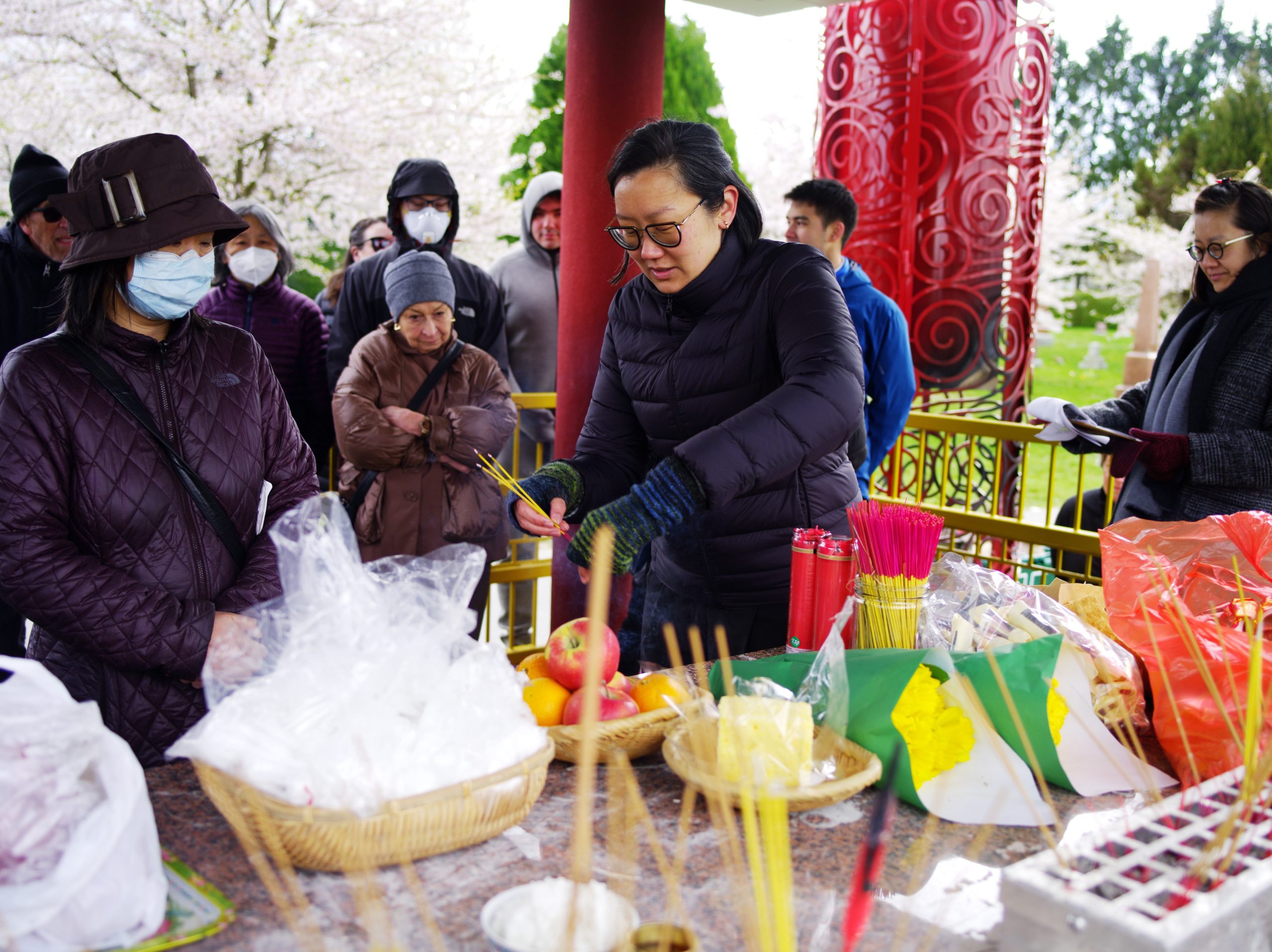 Image Description: Two people stand at the Chinese Pavilion Altar. They are both wearing puffy coats. One person prepares a small bundle of lit incense sticks with a piece of ribbon. The other person watches the lit incense sticks begin to smoke. In the front of the photograph the Altar is bursting with desserts, flowers, incense, bowls of rice and wicker baskets filled with plastic bags. A group of people watch the activity at the Altar from the back of the photograph.