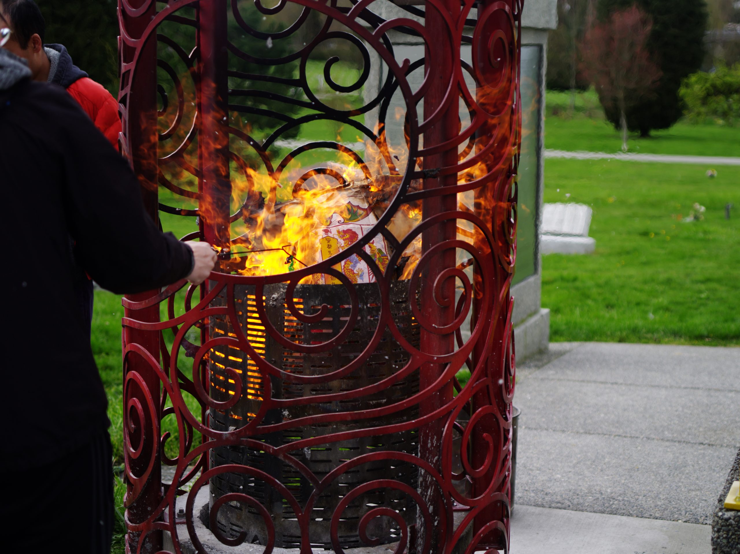 Image Description: A person’s right arm repositions materials with a long thin wire tool inside a fire in the funerary burner at the Chinese Pavilion at Mountain View Cemetery. A decorated paper commemoration is being turned to ash by the heat and the flames. Smoke and ash float above the fire, while waves of heat distort the surrounding image of the photograph. Flames are visible above and through the metal grate that safely holds the offerings provided by community members.
