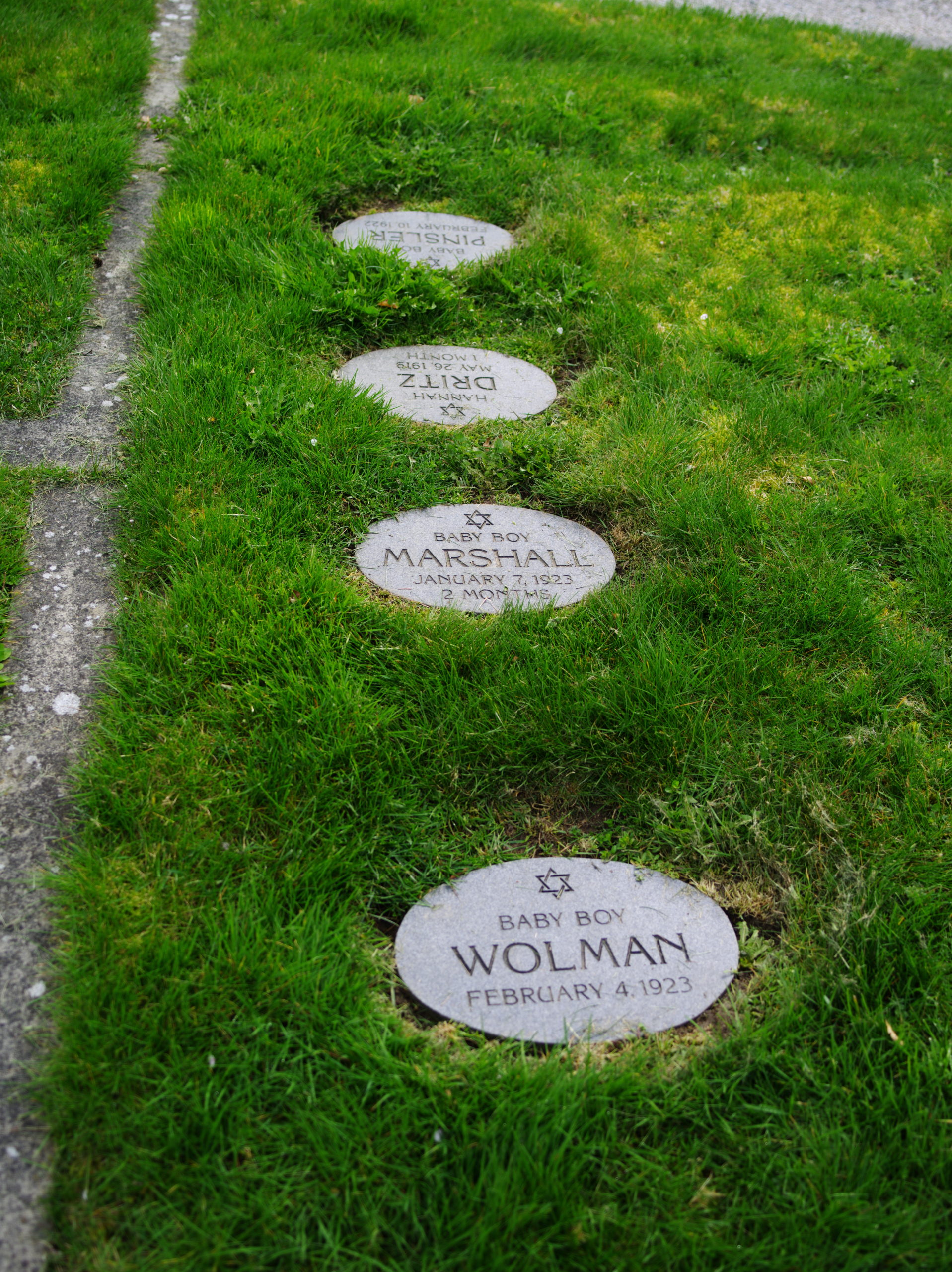 Image Description: Four flat oval head stones are nested in a vertical line into the grassy burial site of the Jewish Cemetery. At the top of each headstone is a six pointed star, also called the Star of David.The stone closest to the camera reads: BABY BOY WOLMAN FEBRUARY 4, 2023 The stone behind this stone in the photograph reads: BABY BOY MARSHALL JANUARY 7, 1923 2 MONTHS The next stone is oriented away from the camera in the photograph, which reads: HANNAH DRITZ MAY 26, 1919 1 MONTH The stone farthest away from the camera in the photograph reads: BABY BOY PINSLER FEBRUARY 10, 1923