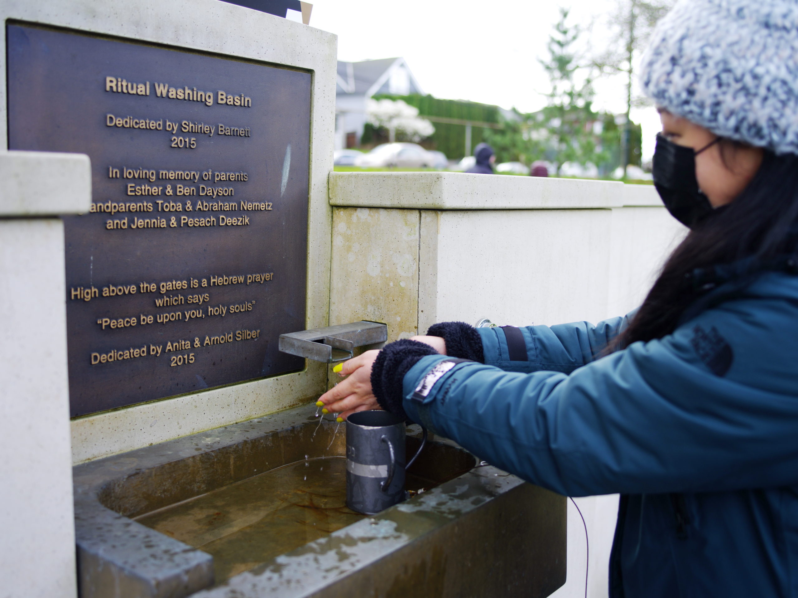 Image Description: A person wearing a heavy winter coat washes their hands in the Ritual Washing Basin. Their long dark hair falls over their left shoulder. They have on a knitted hat and a face mask. Bright neon nail polish decorates their fingers. There is a dedication plaque above the Ritual Washing Basin. The plaque inscription, written in English reads: Ritual Washing Basin Dedicated by Shirley Barnett 2015 In loving Memory of parents Esther & Ben Dayson Grandparents Toba & Abraham Nemetz And Jennia & Pesach Deezik High above the gates is a Hebrew prayer which says “Peace be upon you, holy souls” Dedicated by Anita & Arnold Silber 2015