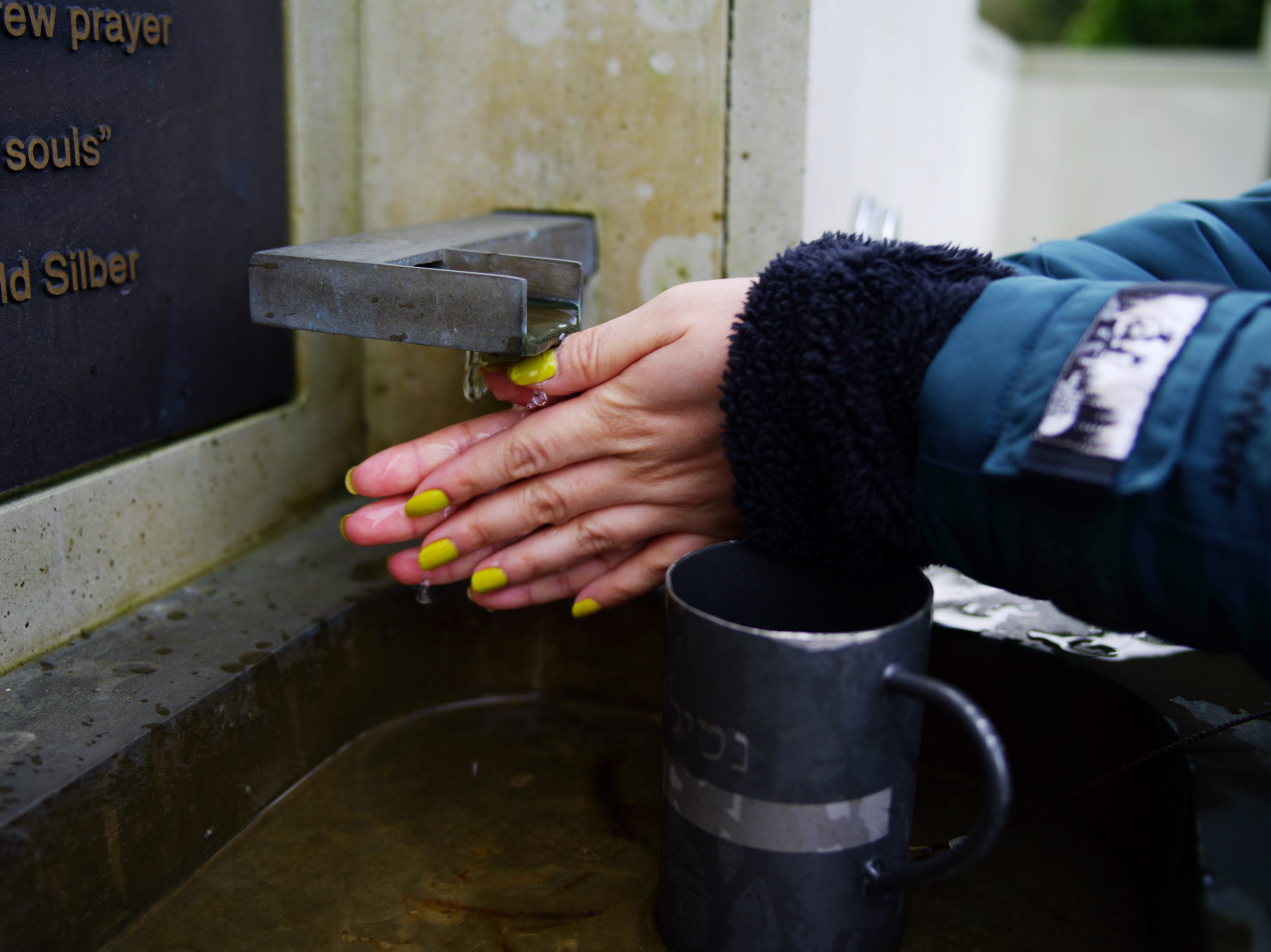 Image Description: Two hands with bright neon nail polish are washed with running water from the metal Ritual Basin. The stream of water is trickling out of a thin rectangular pipe with a 90 bend. A metal cup with Hebrew writing is sitting in the basin, closer to the camera than the two washing hands. The cup has a clip on the bottom attached with a thin metal safety wire.