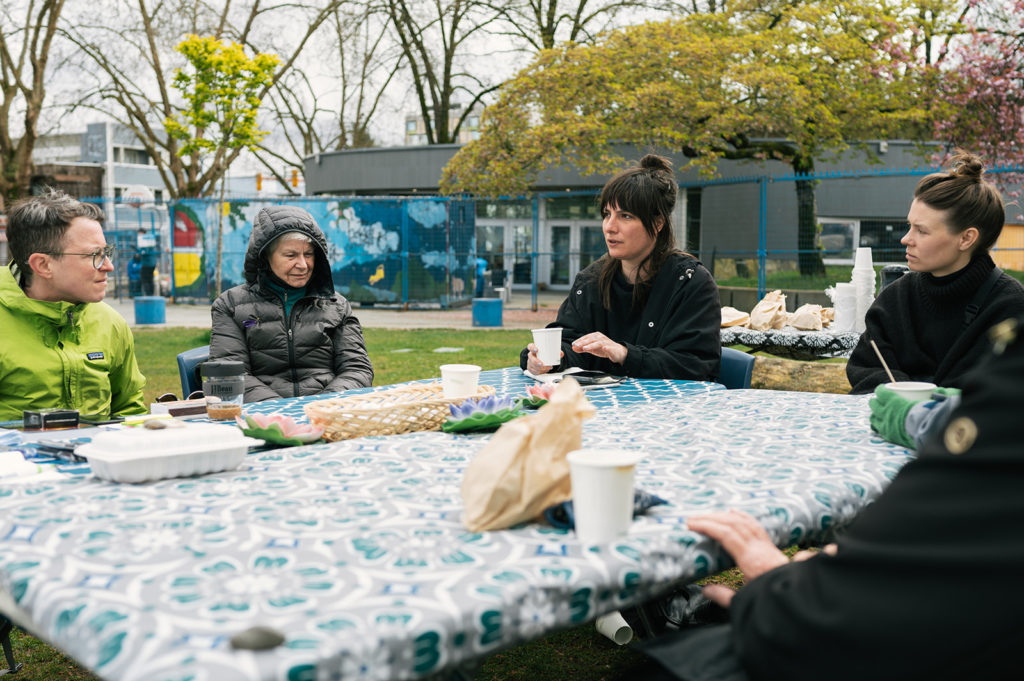 Four people sit at rectangular tables outside at Oppenheimer Park. A fifth and sixth person are outside of the photo. Only their arms and hands are visible. The tables are covered with brightly colored patterned tablecloths. Paper cups and small paper bags are on the table.

A person in the center of the photograph is wearing a dark coat and has their dark hair pulled back away from their face. Their mouth is open, and they are holding their left hand up. The person on the far left of the photograph, and the person on the far right of the photograph are watching them intently. Another person in a puffy jacket with a hood pulled up, is looking down with the reflective expression on their face.
