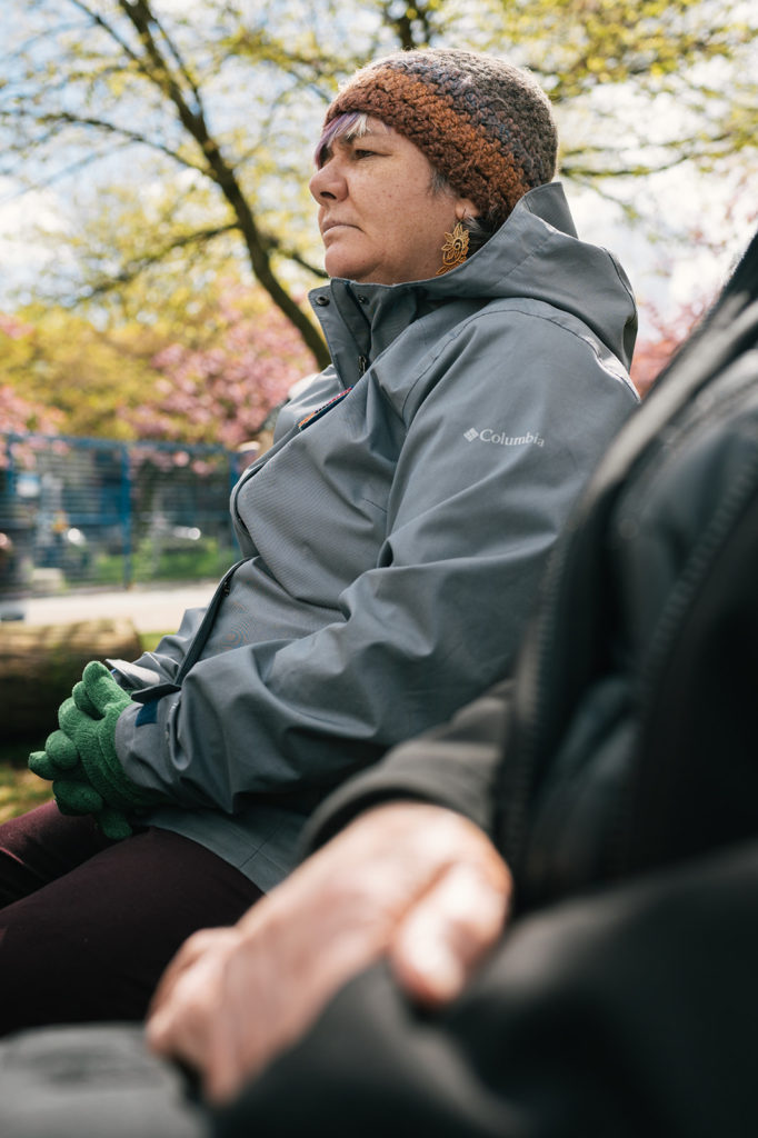 A close-up of a person sitting outside. They are facing the left side of the photograph. Wearing a knitted cap, their pink and purple bangs are pressed close to their forehead. They are wearing a wet weather jacket with a hood, gloves, and dark pants.

The person is wearing earrings made of delicately crocheted lace dyed a warm color. Their fingers are folded together in their lap. 
