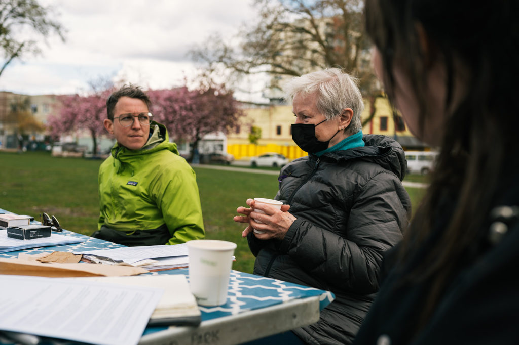 There are three people seated at a table outside. A person in a dark, puffy jacket, holds a white paper cup in their two hands. Their face is covered by a black cloth face mask. Their short white hair is ruffled.

The person to their right looks at them intently. The person on their left, closest to the photographer, is out of focus.
