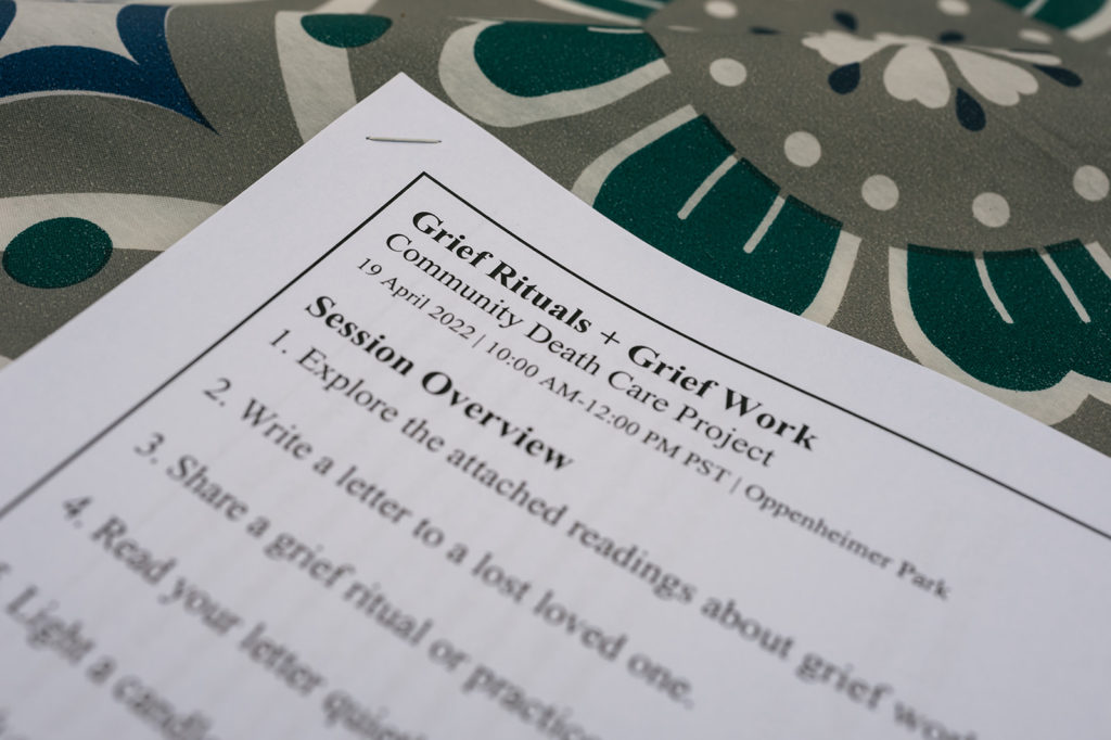 A close up photograph of stapled worksheets.The title reads:
Grief Rituals + Grief Work
The worksheets are on a brightly patterned tablecloth made up of teal, blue, grey, black, and cream coloured floral designs.
