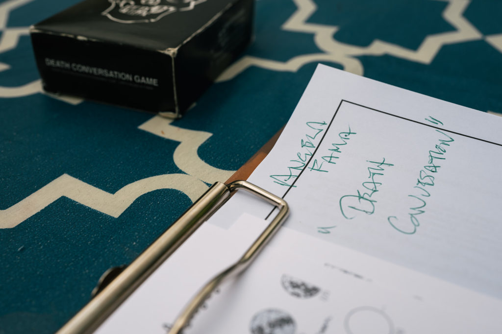 An extreme close up of the top right corner of a clipboard holding hand written and typed notes. 
Handwritten in green ink can be read:
Angela
Fama
“Death 
Conversation”
In the top left corner of the photograph, a black box holding a deck of cards is out of focus. It is labeled “DEATH CONVERSATION GAME” in white ink.
