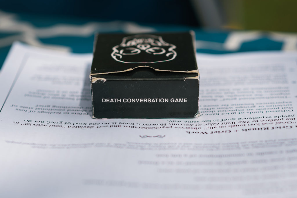 A glossy black box holding a deck of cards sits on top of a white piece of paper filled with typed words in English. The edges of the glossy black box are worn and raggety. 

Printed on the long narrow edge of the box facing the camera reads:

DEATH CONVERSATION GAME

In white ink. 

An animated outline of a skull is out of focus but visible on the top of the black box, facing away from the camera towards the top of the photograph.
